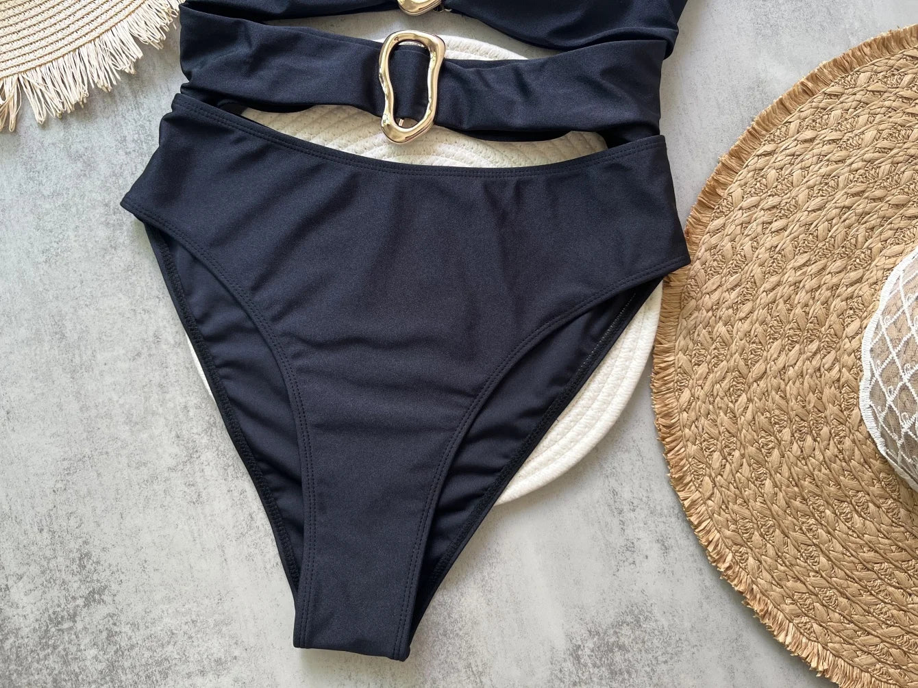 Black swimsuit with a gold buckle on a light gray surface with straw hats.
